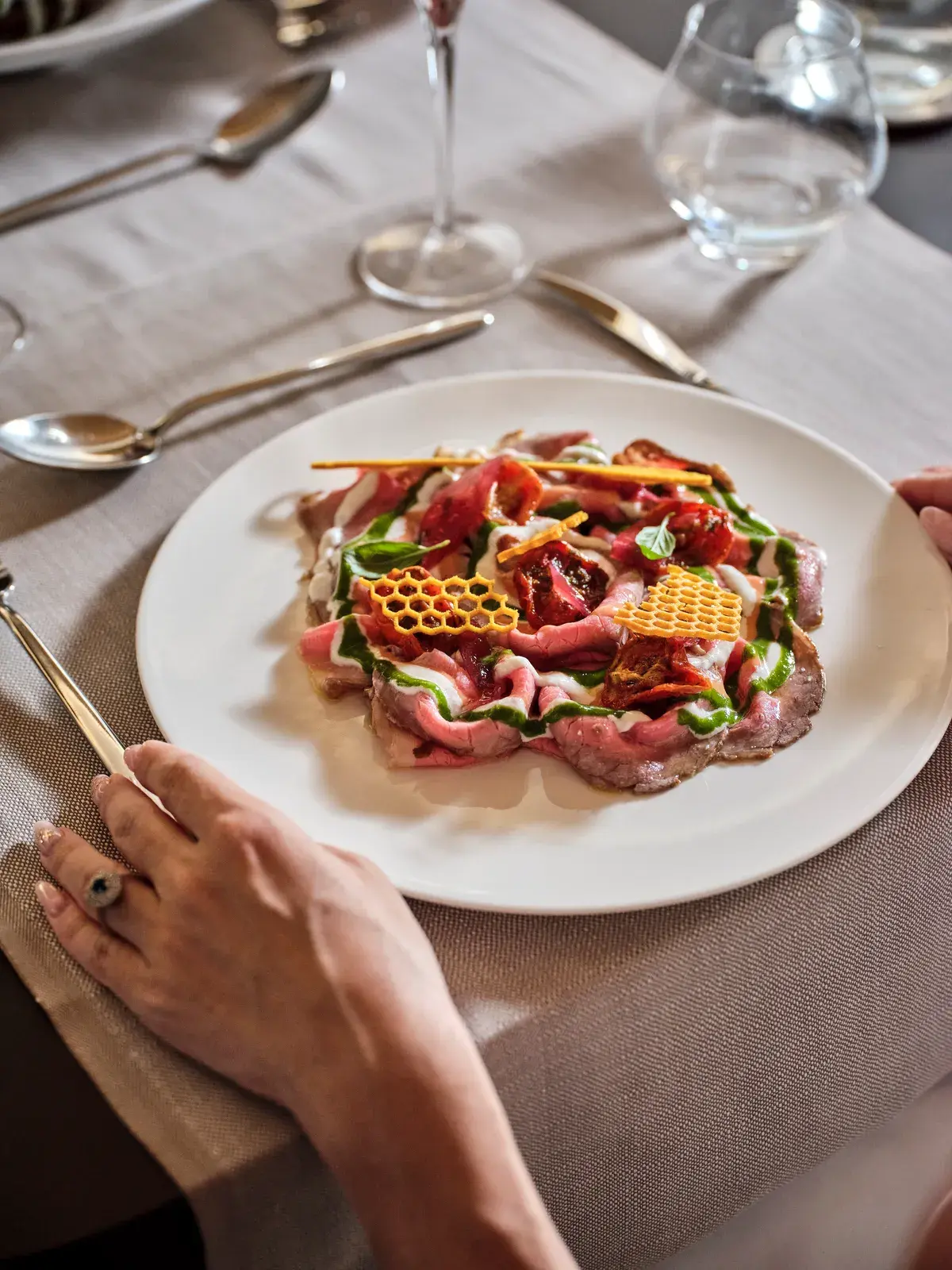 Roastbeef alla Pugliese affettato sottile con pomodorini confit, gel verde di prezzemolo e cialde a nido d'ape; le mani di un ospite a tavola apparecchiata