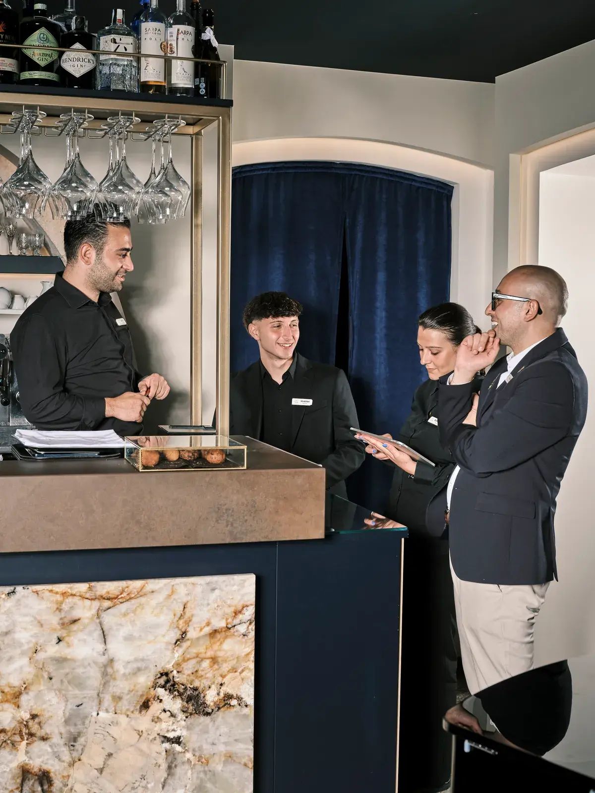 Lo staff in nero sorride con un ospite all'angolo bar, tra bottiglie illuminate e base in marmo venato