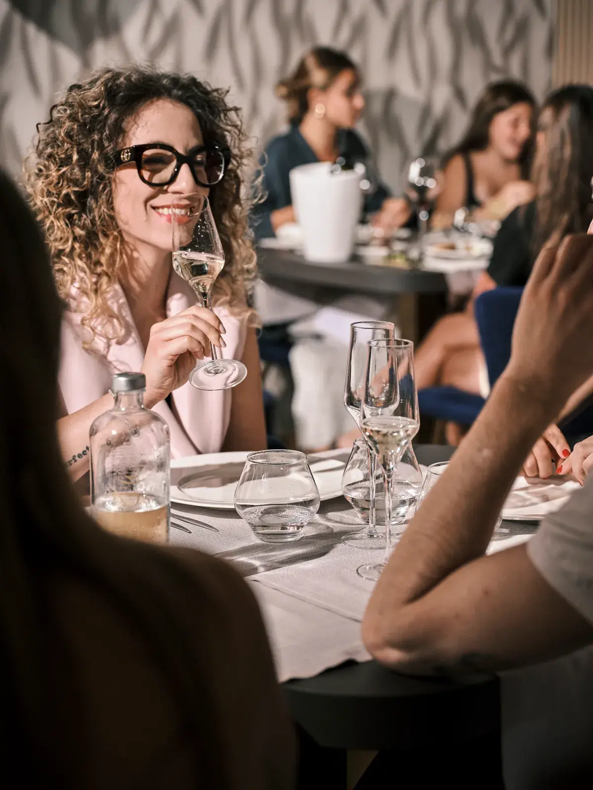 Donna sorridente con occhiali assapora un calice di bollicine a tavola, sala calda con altri ospiti sullo sfondo